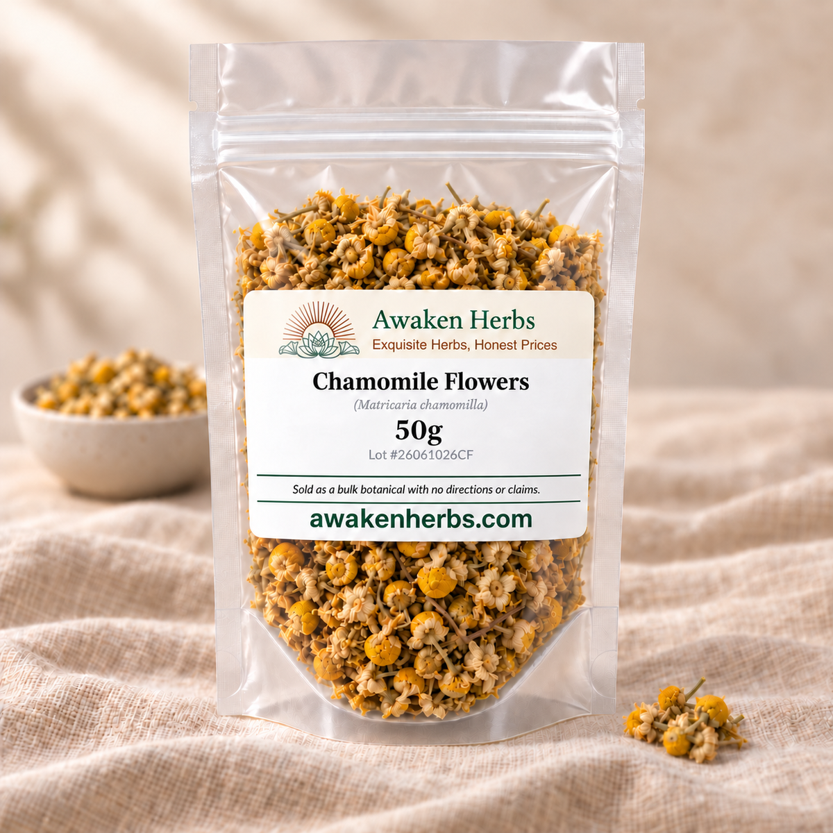 Awaken Herbs Chamomile Flowers 50g clear pouch with an ivory label, beside a softly blurred bowl of chamomile on beige linen.