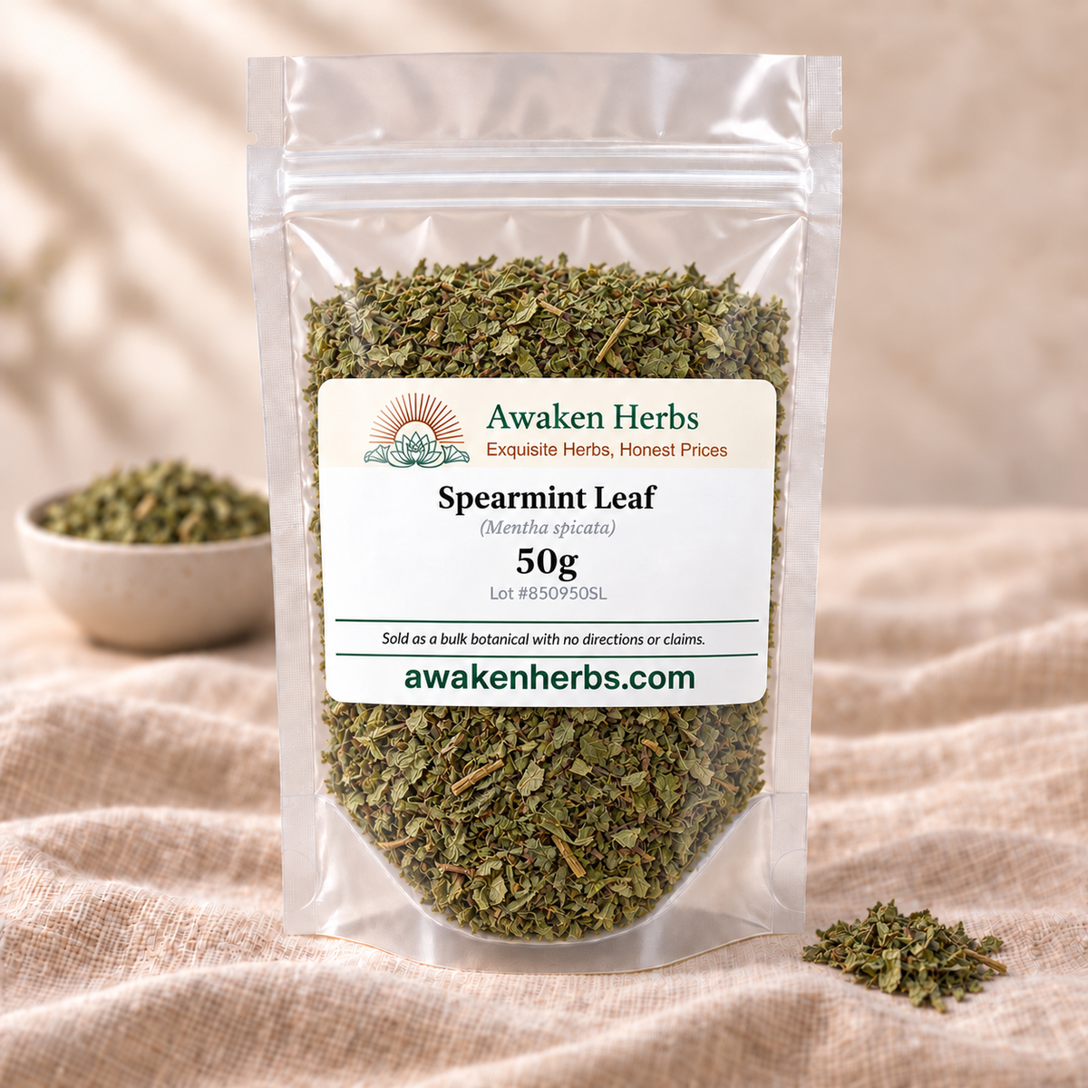 Awaken Herbs Spearmint Leaf 50g clear pouch with an ivory label on beige linen, beside a softly blurred bowl of the same dried botanical.